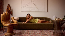 Woman in yellow dress reclines on olive green sofa in styled interior with sculptural wooden furniture.