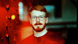 Man wearing glasses and red shirt photographed through rain-covered glass with warm and cool color cast.