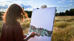 Artist painting a landscape scene on canvas outdoors in an open field during golden hour.