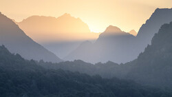 Sun rising between forested mountain peaks shrouded in layered mist and haze.
