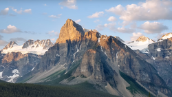 Dramatic mountain peak with vertical columnar rock formations, snow patches, and forested valley below under partly cloudy sky.