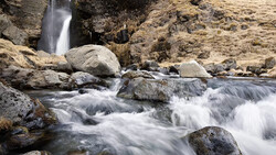 Mountain stream with cascading white water flowing around moss-covered rocks and boulders, with a waterfall visible in the background.