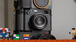 Stack of vintage cameras including a Ricoh GR film camera and Leica lens with pixel art figures and colored film boxes in foreground.