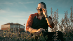 Man in orange shirt and sunglasses crouching in a field during golden hour light.