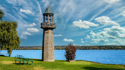 Stone lighthouse with green observation deck beside calm blue water and grassy shoreline.