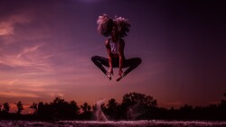 Person with curly hair jumping above water during purple sunset, captured mid-air in silhouette.