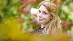 Young woman with long blonde hair looking over her shoulder in an outdoor garden setting.
