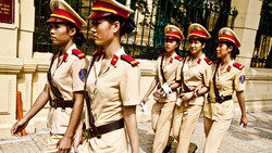 Five uniformed personnel in beige military-style uniforms with red armbands and caps standing in formation outdoors.