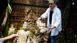 Man in light blue shirt gesturing toward stone sculptures in an overgrown courtyard with carved relief panels.