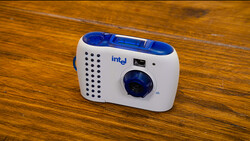 Intel-branded compact point-and-shoot camera with white body, blue accents, and perforated speaker grille on wooden surface.