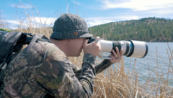 Wildlife photographer with telephoto lens capturing images by a forested lake shore.