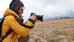 Photographer in yellow jacket and hood holding DSLR camera while crouching in open field.
