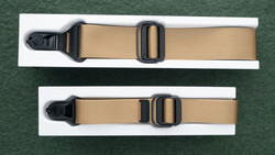Two tan webbing straps with black plastic side-release buckles displayed on white cards against a green background.