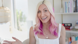 Woman with blonde and pink ombre hair smiling at camera in bright interior space.