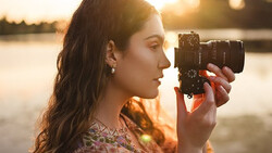 Woman holding a mirrorless camera to her face during golden hour, silhouetted against a body of water.