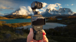 DJI Osmo Pocket gimbal camera held in hand against snowy mountain peaks and turquoise lake landscape.