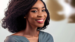 Woman with curly dark hair smiling at camera, wearing off-shoulder blue-gray top.