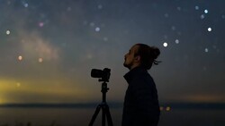 Photographer silhouetted against twilight sky, aiming camera on tripod toward starry horizon.