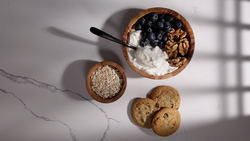 Overhead flat lay of breakfast ingredients in wooden bowls: yogurt with blueberries and granola, oats, and cookies on marble surface.