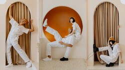 Three people in white clothing posing dynamically in arched doorways with tan curtains and an orange circular backdrop.