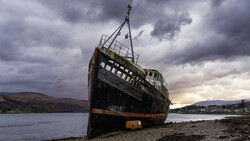 Abandoned ship beached on rocky shore with dramatic overcast sky and mountains in background.