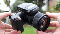 Sony camera with flash and telephoto lens held in both hands against outdoor background.