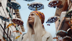 Woman with long platinum blonde hair and gold headwrap gazing upward surrounded by hanging decorative UFO sculptures.