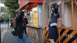 Two students in school uniforms posing playfully against urban storefronts and metal structures.