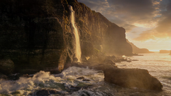 Waterfall cascading down a steep coastal cliff face during golden hour, with waves crashing on rocks below.
