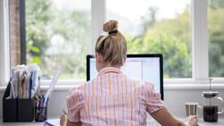 Woman working at desktop computer near bright windows with natural outdoor light streaming in.