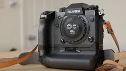 Fujifilm X-H1 mirrorless camera with battery grip and brown leather strap on wooden surface.