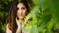 Woman with long brown hair posing among green foliage with soft natural lighting.