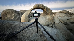 Smartphone mounted on tripod framing a natural rock arch against cloudy sky.