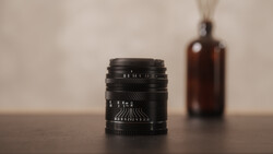 Black vintage lens with depth of field markings sits on wooden surface with brown glass bottle blurred in background.