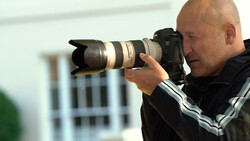 Photographer holding a DSLR camera with a large telephoto lens, composing a shot outdoors.