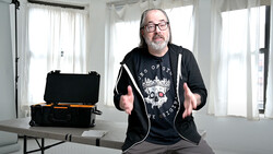 Man in glasses and black hoodie gestures while speaking in bright studio with equipment case on table.