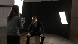 Photographer operating camera while interviewing man in suit in professional studio setup with large soft box lighting.