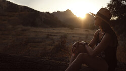 Woman in hat sitting outdoors at golden hour, silhouetted against sunset landscape.