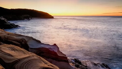 Rocky coastline at sunset with layered sandstone formations and calm ocean waters.