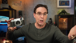 Man holding a vintage rangefinder camera while speaking to camera in a studio setting.