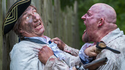 Two men in period costume engaged in a physical altercation near a wooden structure.