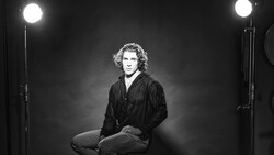 Man with curly hair sitting in studio lighting, wearing dark pinstriped shirt against dark background.
