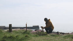 Photographer crouching with tripod-mounted camera on a pebbly beach overlooking water.