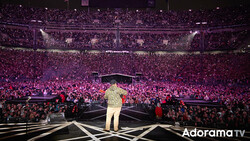 Performer in cream-colored suit standing on stage facing massive crowd illuminated by pink lighting in large arena.