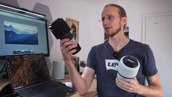 Man holding a cylindrical black device while gesturing toward a laptop displaying landscape photography.