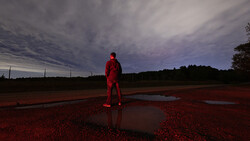 Figure in red clothing standing alone on wet red earth with water puddles under dramatic twilight sky.