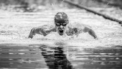 Competitive swimmer performing butterfly stroke in rain, captured mid-stroke with water splashing around their face and goggles.