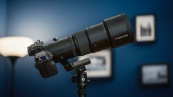 Fujifilm telephoto lens mounted on tripod against blue studio backdrop with out-of-focus frames on wall.