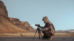 Photographer crouching beside a tripod-mounted camera overlooking an arid desert landscape with rock formations.