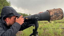 Photographer using a telephoto lens with camouflage barrel cover mounted on a tripod in an open field.